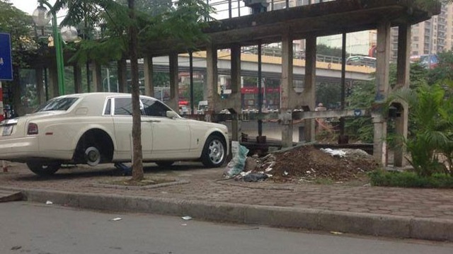 
Một chiếc Rolls Royce Phantom nằm lặng lẽ chờ thay bánh trên vỉa hè Hà Nội, bên cạnh là bãi rác.
